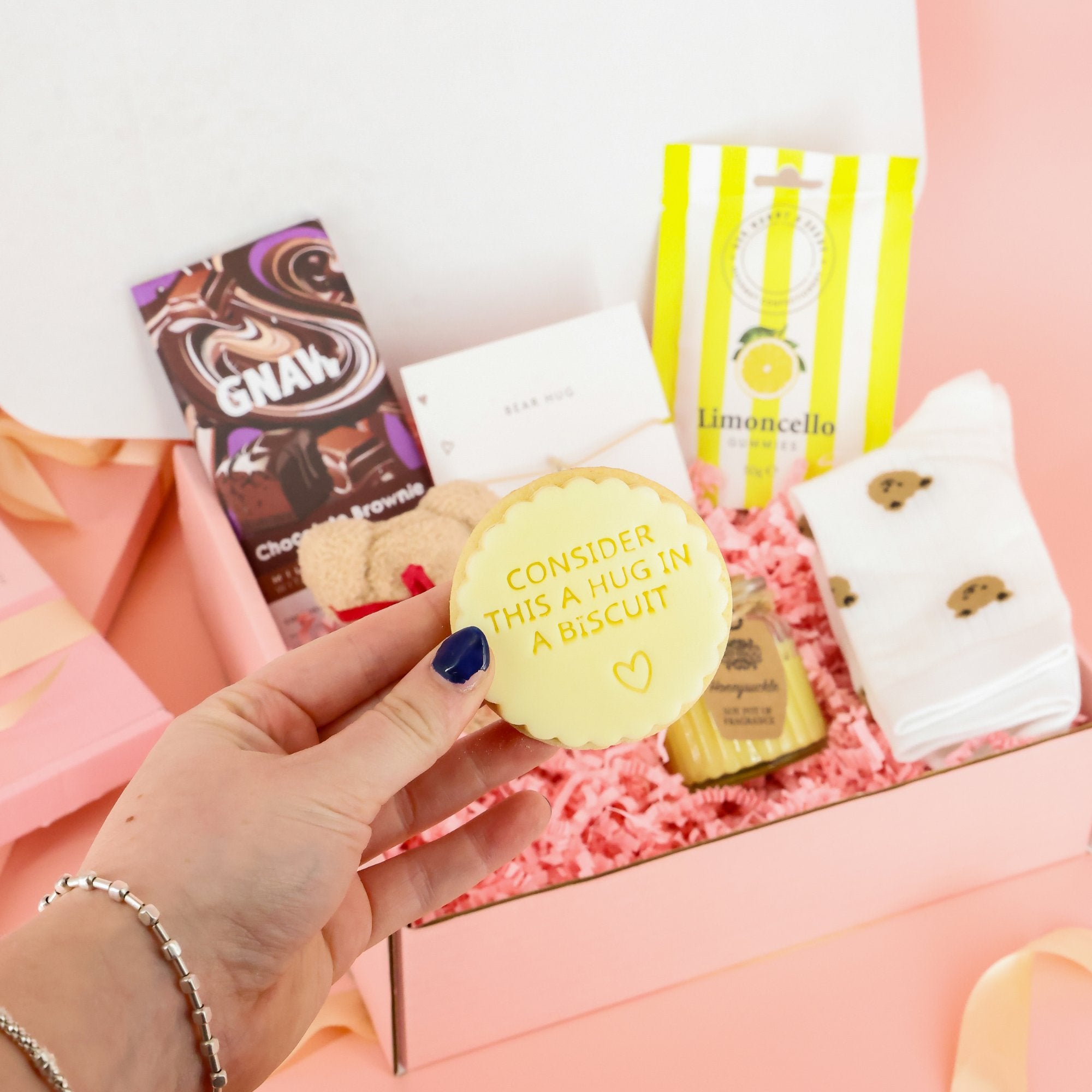 Close up of a hand holding a "Consider this a hug in a biscuit" iced cookie from a thinking of you gift box and virtual hug care package.