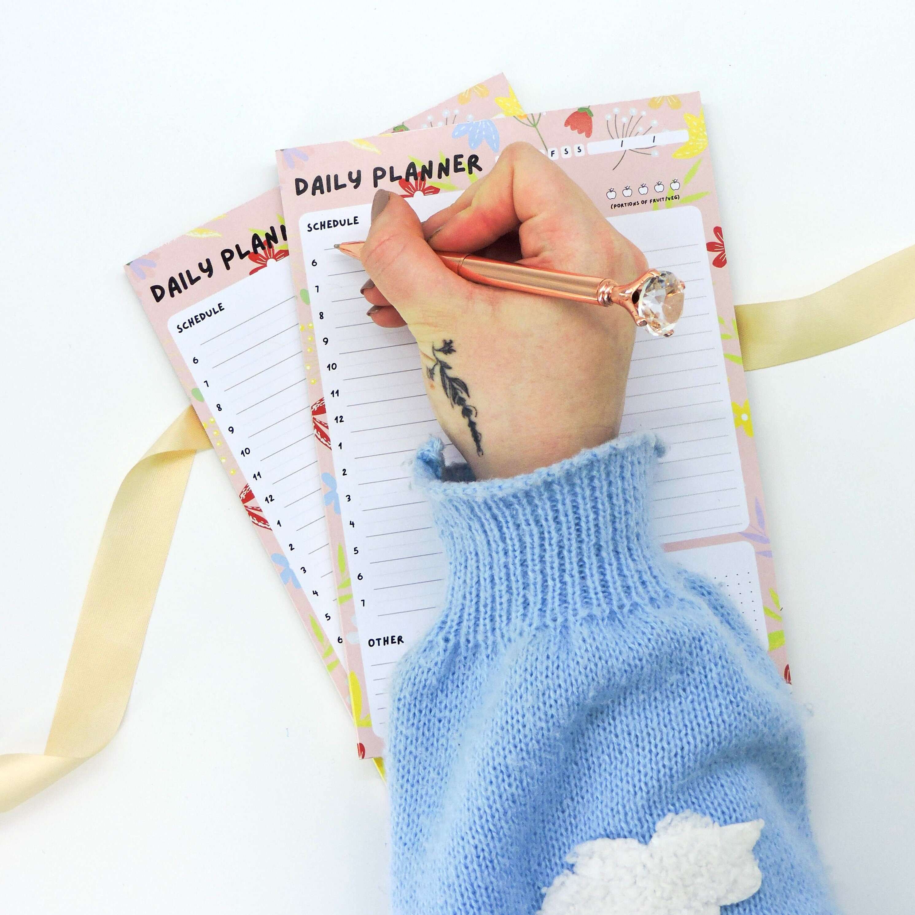 Daily Planner A5 Deskpad in use, with a hand holding a pen over its floral layout, featuring sections for scheduling and to-do lists.