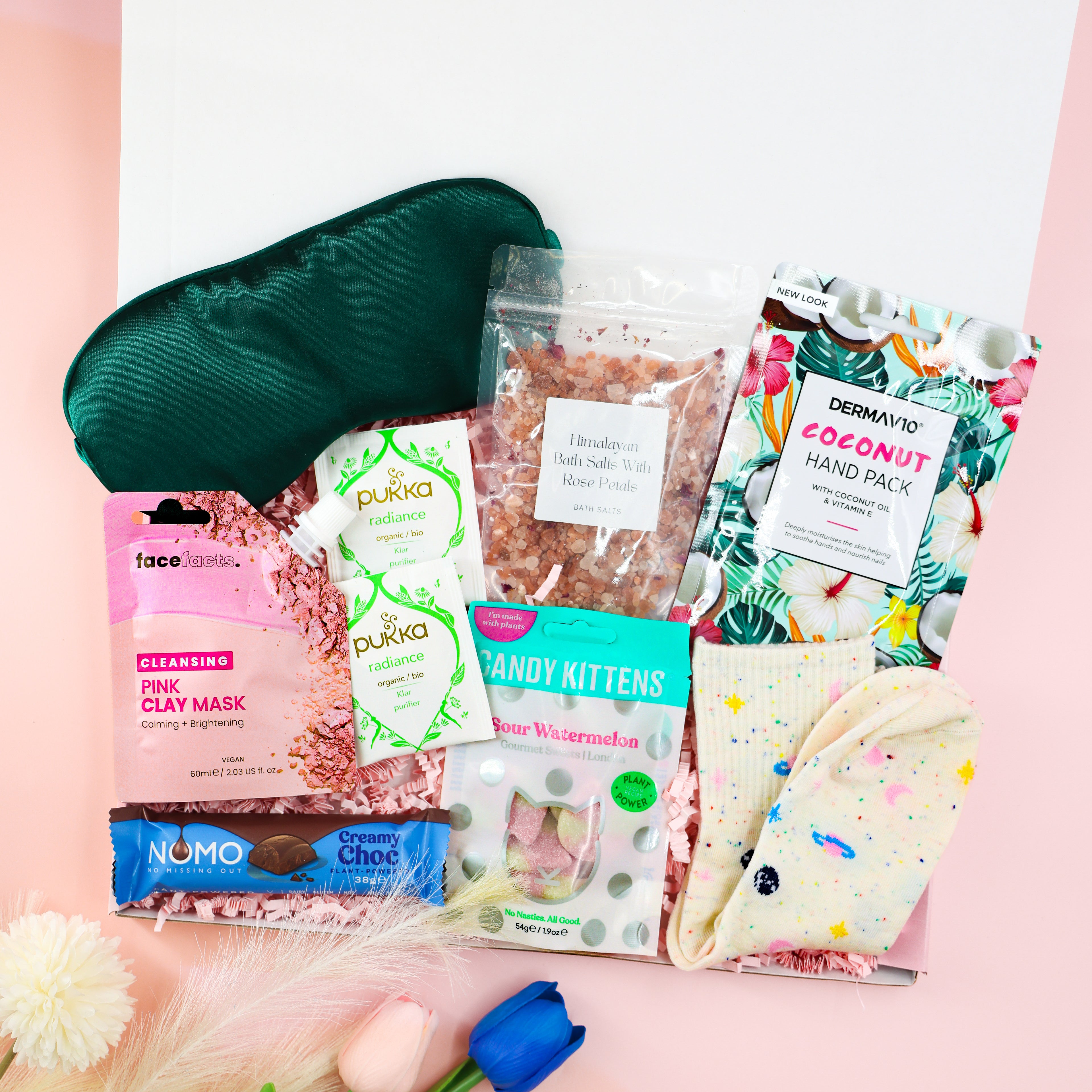 Feel-Good Care Package gift box for her with green satin sleep mask, Himalayan bath salts, coconut hand mask, pink clay mask, Pukka tea, vegan sweets and cosy socks.