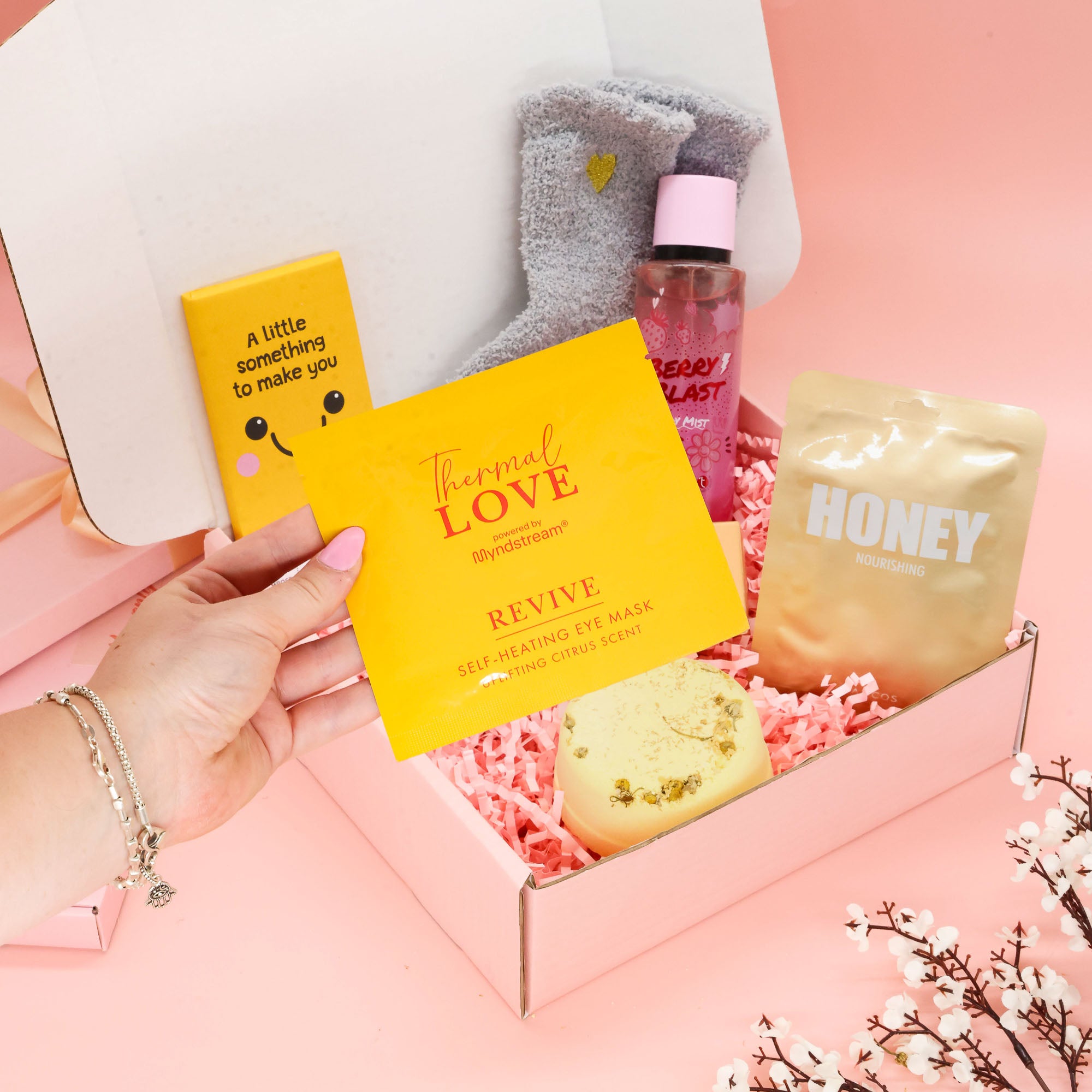 A gift box set for self-care and relaxation, featuring a hand holding a yellow Thermal Love eye mask box, alongside a honey packet, a pink bath bomb bottle, and gray socks inside a pink box on a pink background.