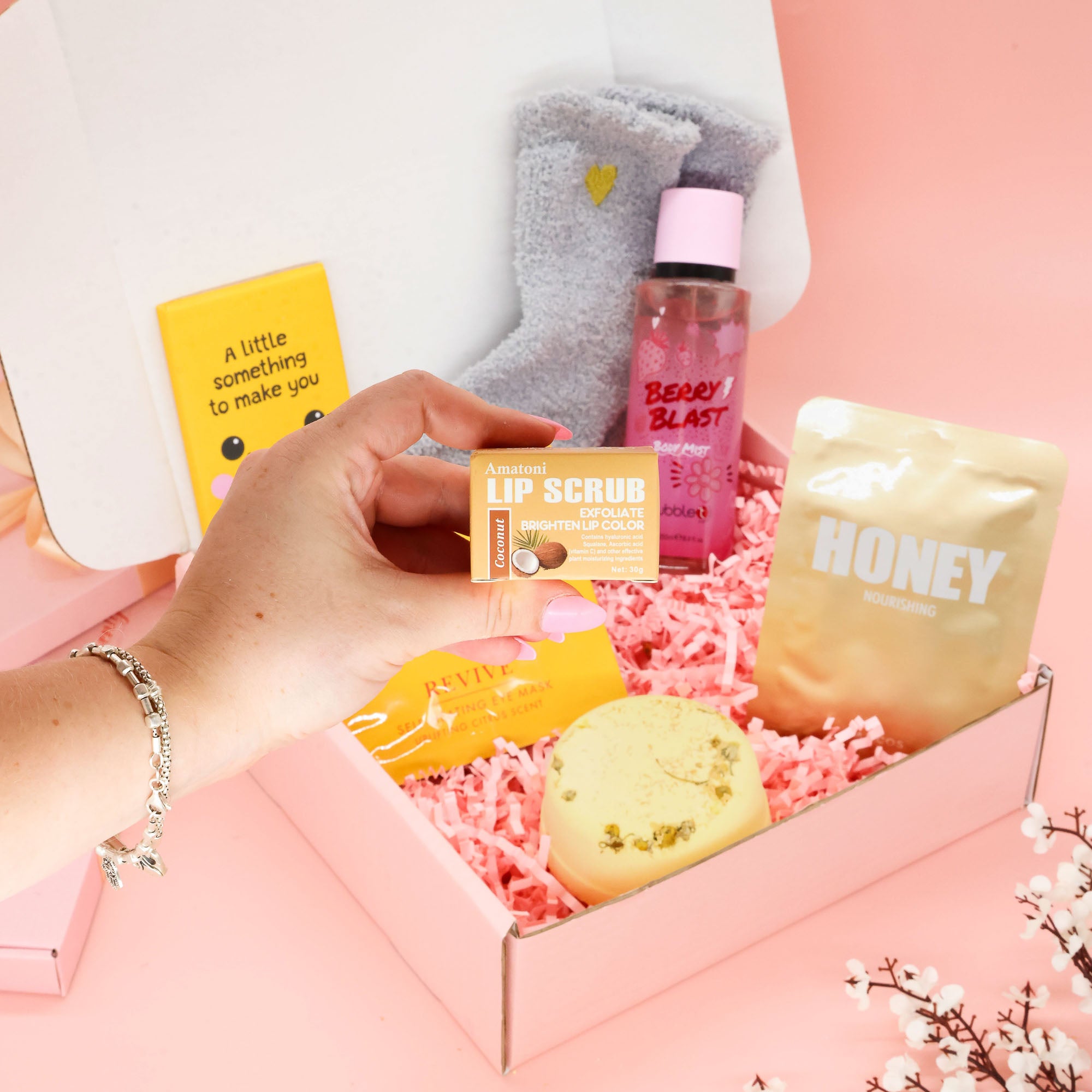 A gift box set containing a yellow lip scrub, a bubble t berry mist bottle, a LAPCOS honey face mask, a grey heart socks, and a card, arranged in pink paper shred inside a pink box, with a hand holding the lip scrub.