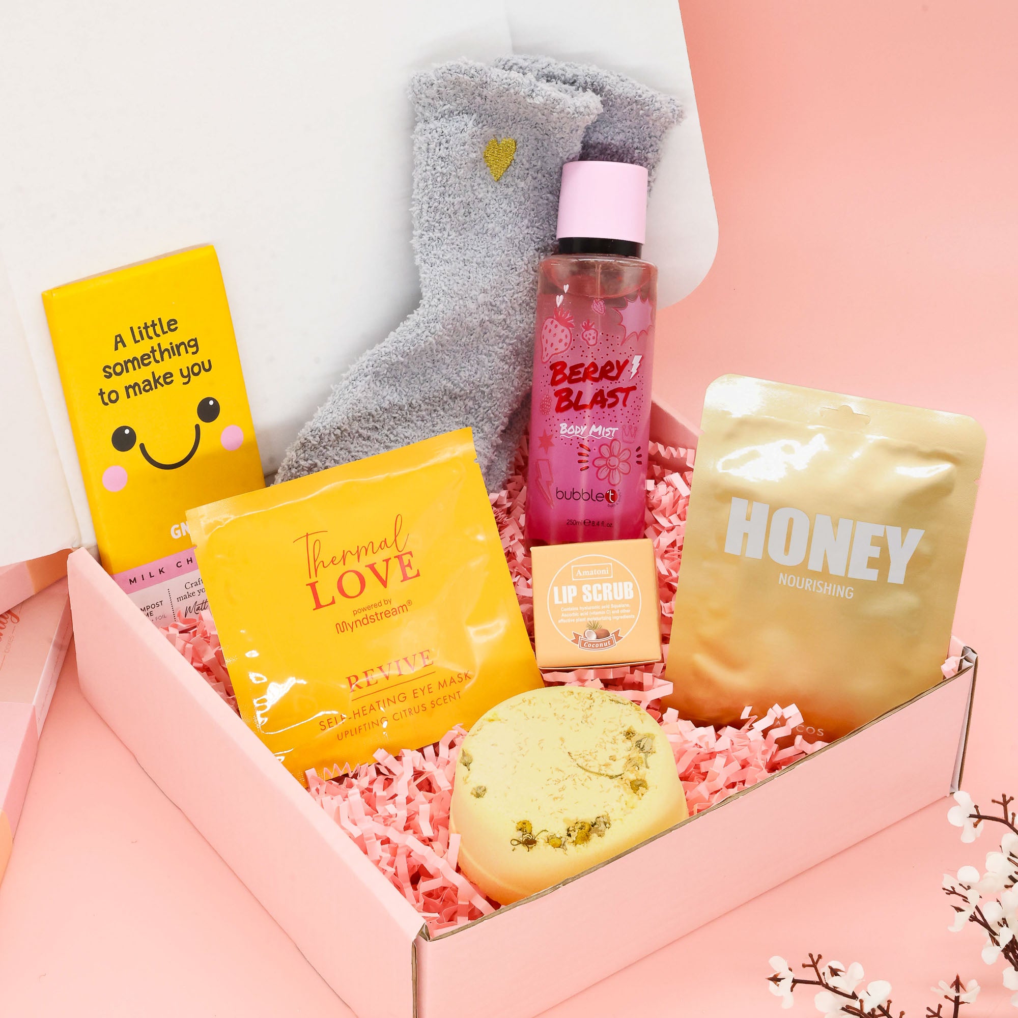 Overhead view of a "Little Box of Sunshine" care package featuring yellow-themed self-care items including a Korean Lapcos sheet mask, fluffy heart socks, bubble t berry blast body mist, and a bath bomb on pink shredded paper.