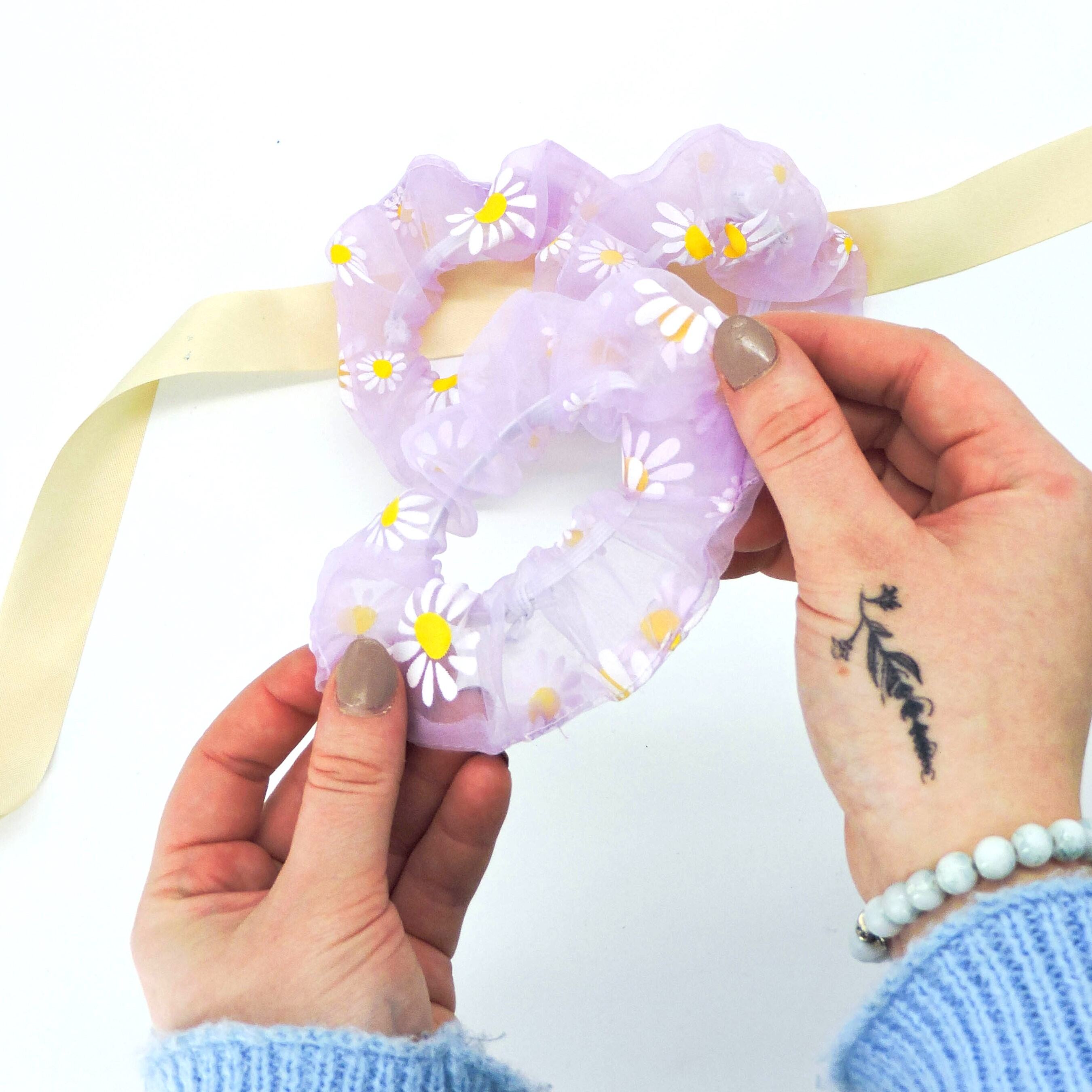 Hand holding Daisy Hair Scrunchie - Purple, featuring white daisies on sheer organza fabric, perfect for styling elegant ponytails or updos.