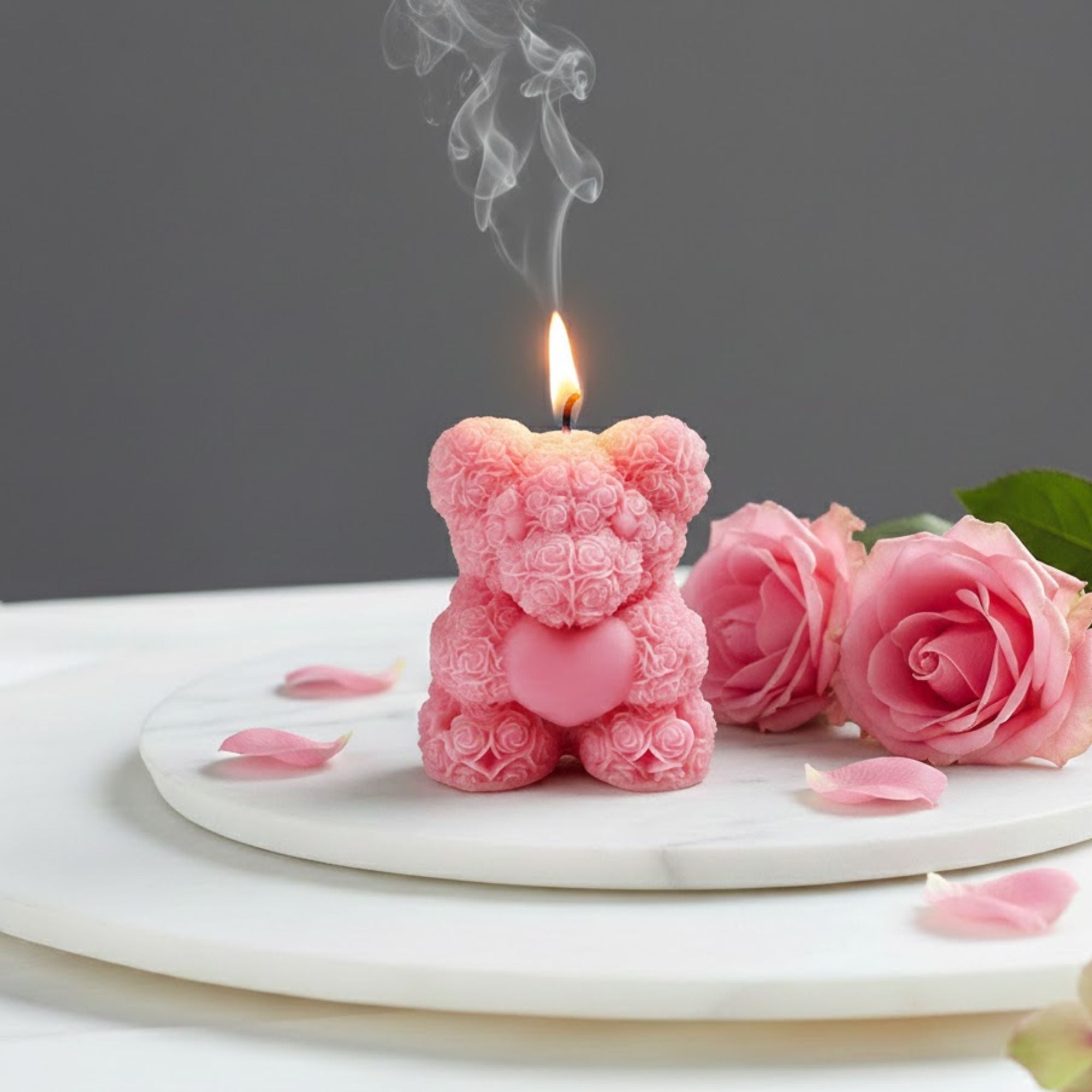 Lit pink rose bear scented candle shaped like a teddy bear made of roses, displayed on a marble tray with fresh pink rose petals for a romantic gift setting.