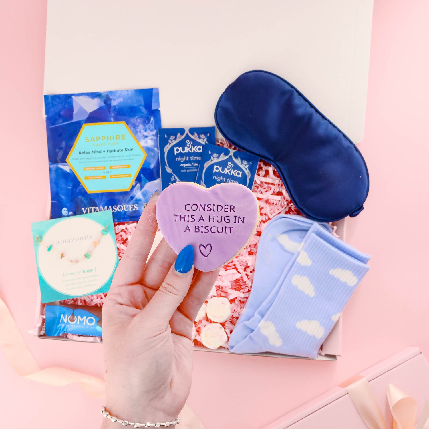 Thoughtful self care gift box for her with pampering treats, herbal teas, cloud socks, satin sleep mask, and a heart-shaped “hug in a biscuit” cookie.