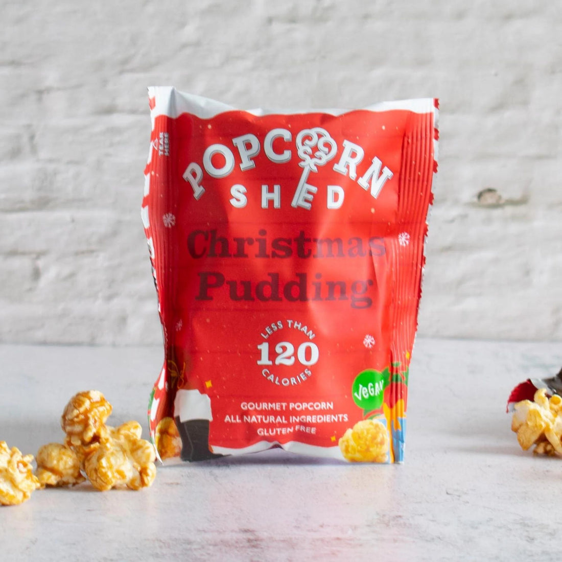 Christmas Pudding Popcorn, a festive snack blend of popped corn, sugar, and spices, showcasing its label and a pile of popcorn on a white surface.