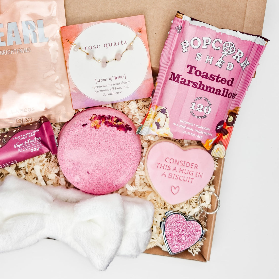 A gift box for her with a LAPCOS PEARL brightening face mask, rose quartz bracelet symbolizing love, a heart-shaped jewel keyring mirror, and toasted marshmallow gourmet popcorn.