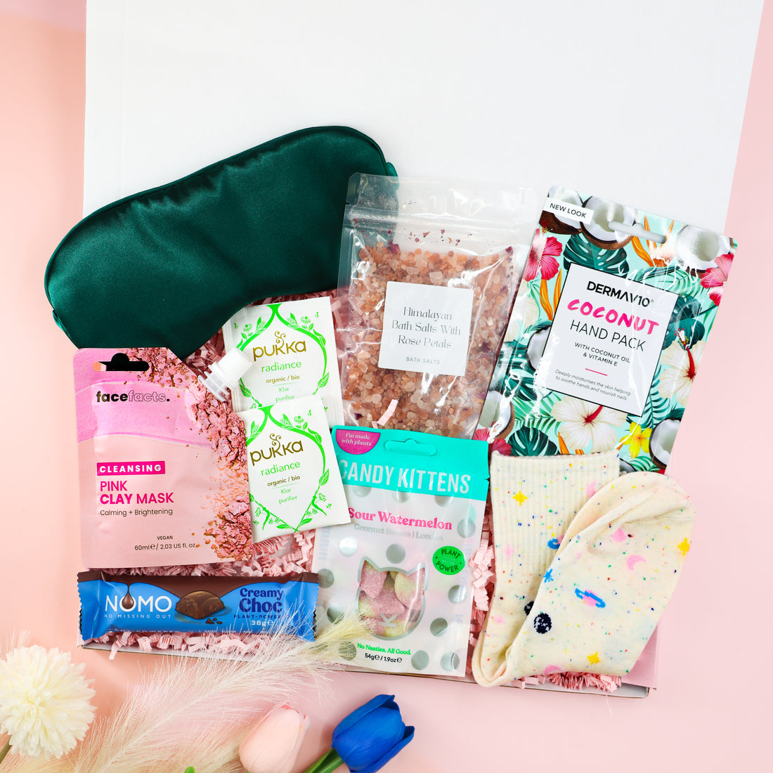 Feel-Good Care Package gift box for her with green satin sleep mask, Himalayan bath salts, coconut hand mask, pink clay mask, Pukka tea, vegan sweets and cosy socks.