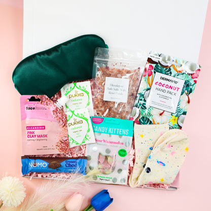 Feel-Good Care Package gift box for her with green satin sleep mask, Himalayan bath salts, coconut hand mask, pink clay mask, Pukka tea, vegan sweets and cosy socks.
