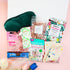 Feel-Good Care Package gift box for her with green satin sleep mask, Himalayan bath salts, coconut hand mask, pink clay mask, Pukka tea, vegan sweets and cosy socks.