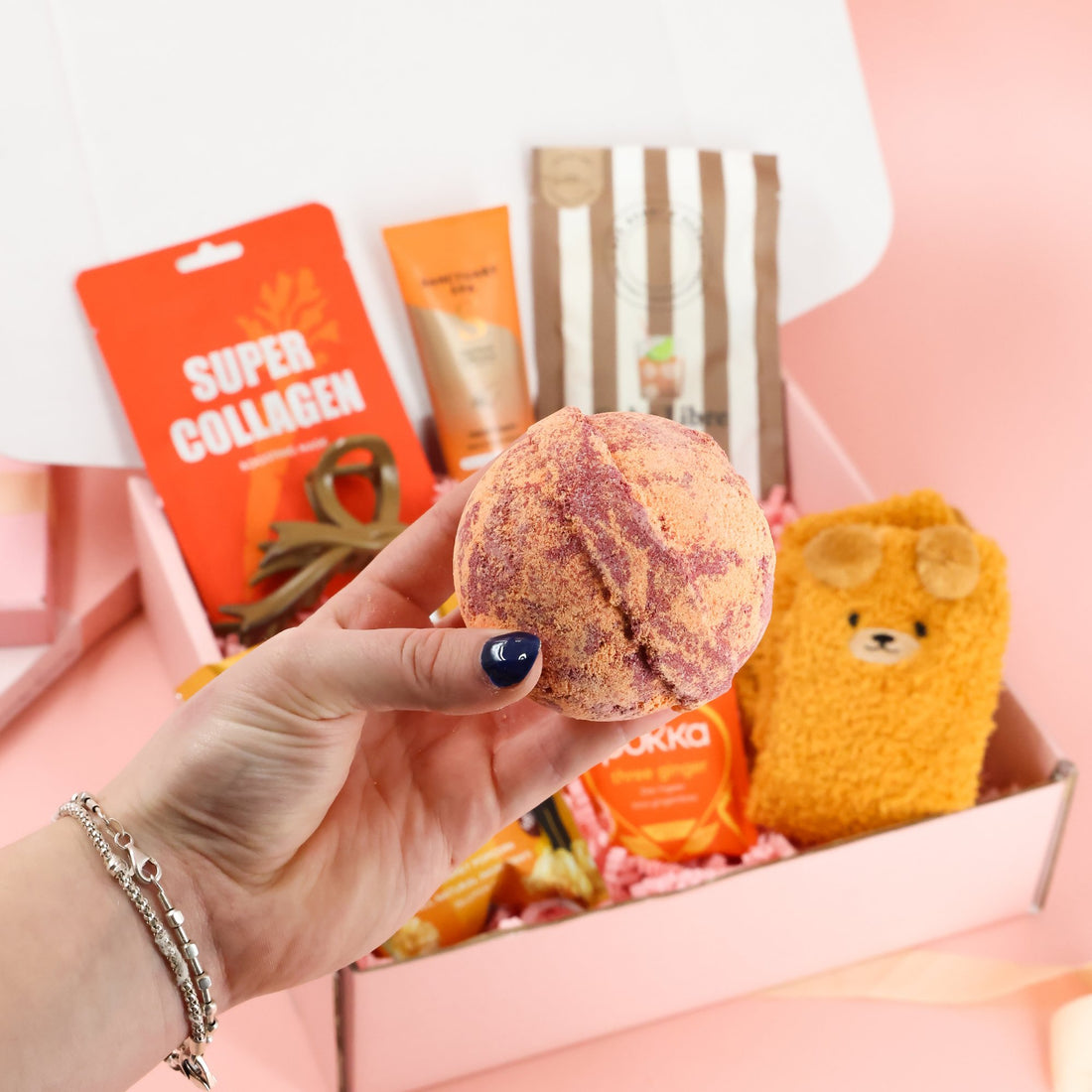 A hand holding a vibrant pink and orange swirling bath bomb in front of a "Hug in a Box" care package containing fuzzy bear socks, herbal tea, and spa treats.