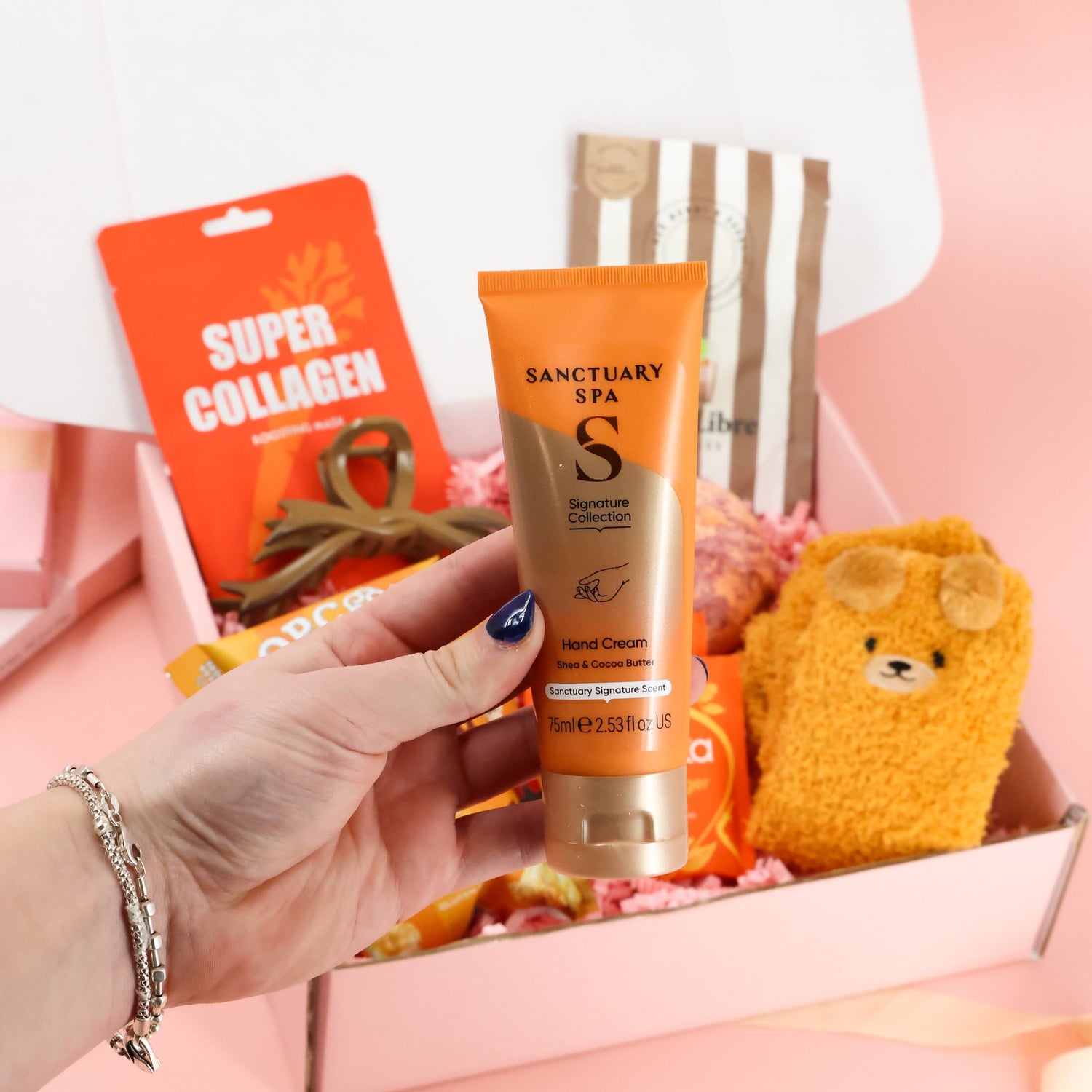 Close up of a tube of Sanctuary Spa Hand Cream being held over an orange themed self care gift box featuring a collagen face mask and cozy accessories.
