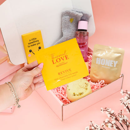 A gift box set for self-care and relaxation, featuring a hand holding a yellow Thermal Love eye mask box, alongside a honey packet, a pink bath bomb bottle, and gray socks inside a pink box on a pink background.