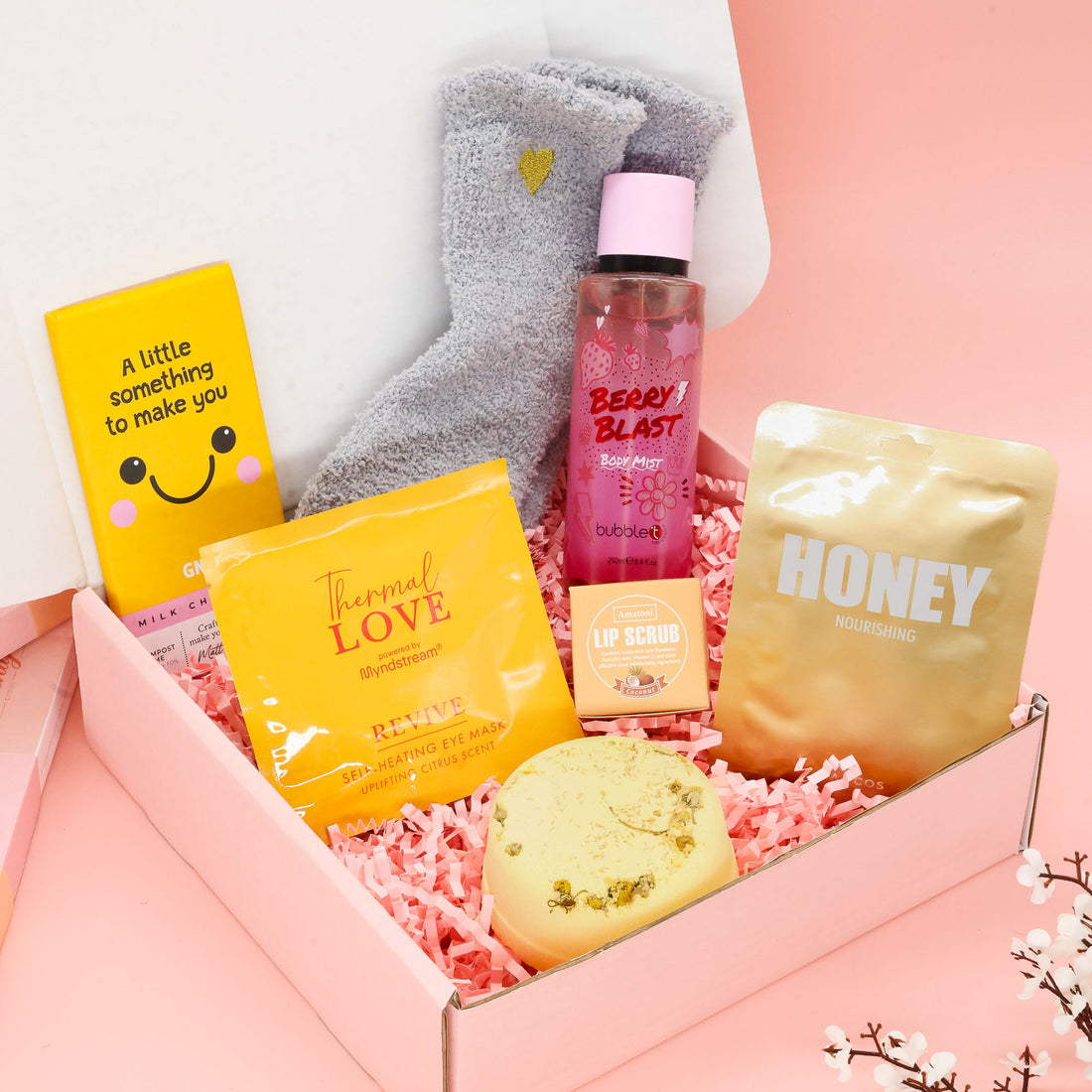 Overhead view of a "Little Box of Sunshine" care package featuring yellow-themed self-care items including a Korean Lapcos sheet mask, fluffy heart socks, bubble t berry blast body mist, and a bath bomb on pink shredded paper.