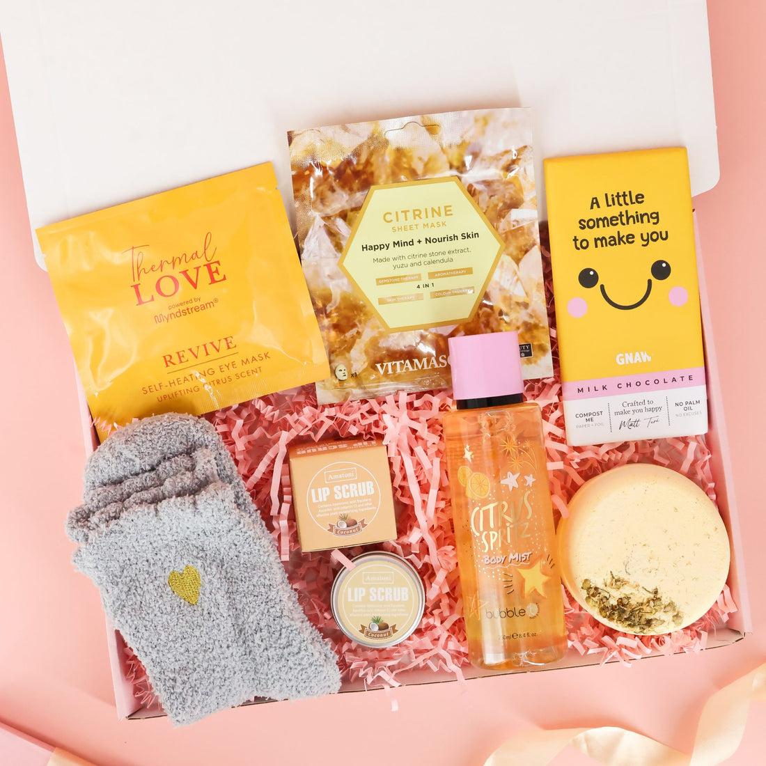 Overhead view of a "Little Box of Sunshine" care package featuring yellow-themed self-care items including a citrine sheet mask, fluffy heart socks, citrus body mist, and a bath bomb on pink shredded paper.