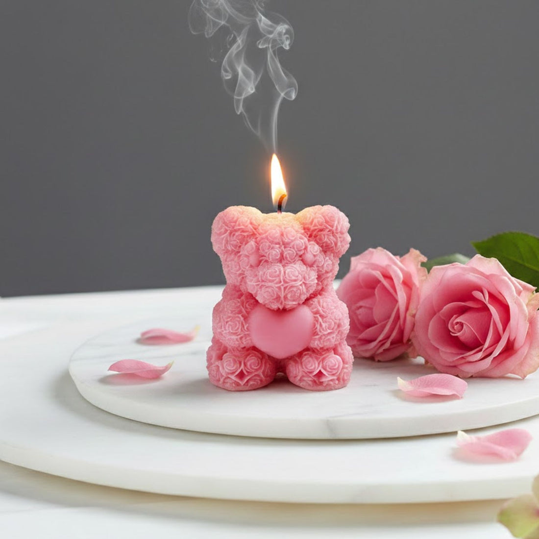 Lit pink rose bear scented candle shaped like a teddy bear made of roses, displayed on a marble tray with fresh pink rose petals for a romantic gift setting.