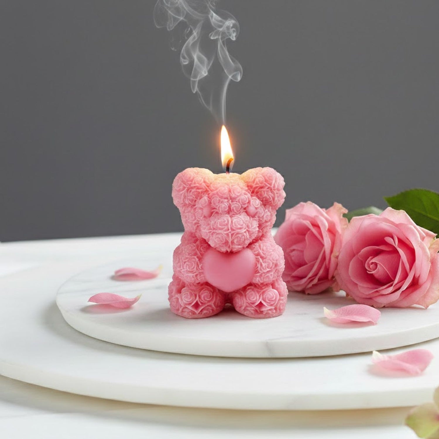 Lit pink rose bear scented candle shaped like a teddy bear made of roses, displayed on a marble tray with fresh pink rose petals for a romantic gift setting.