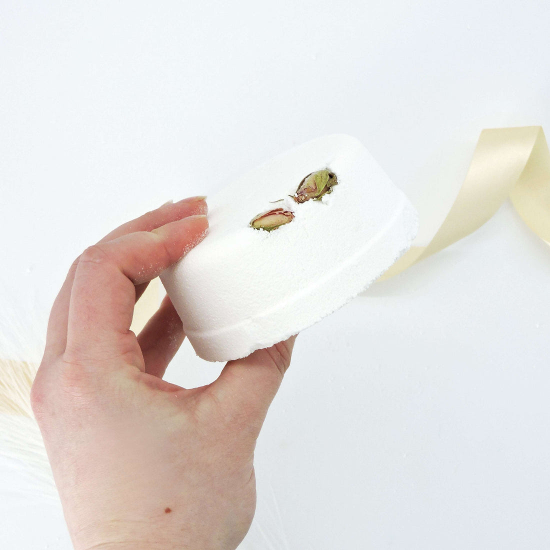 Hand holding the Rose &amp; Ylang Ylang Bath Bomb, a round white object topped with pistachios, designed to soothe and relax with Passion Fashion scent.