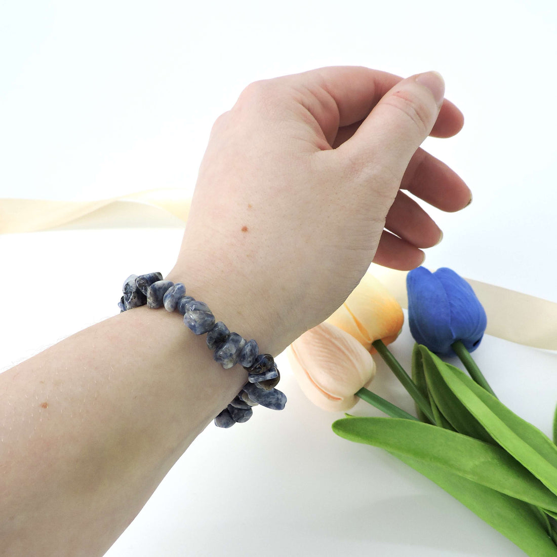 Person wearing the Sodalite Crystal Chip Bracelet, a fashion accessory promoting calmness and intuition, on their wrist amidst a backdrop of blue flowers.