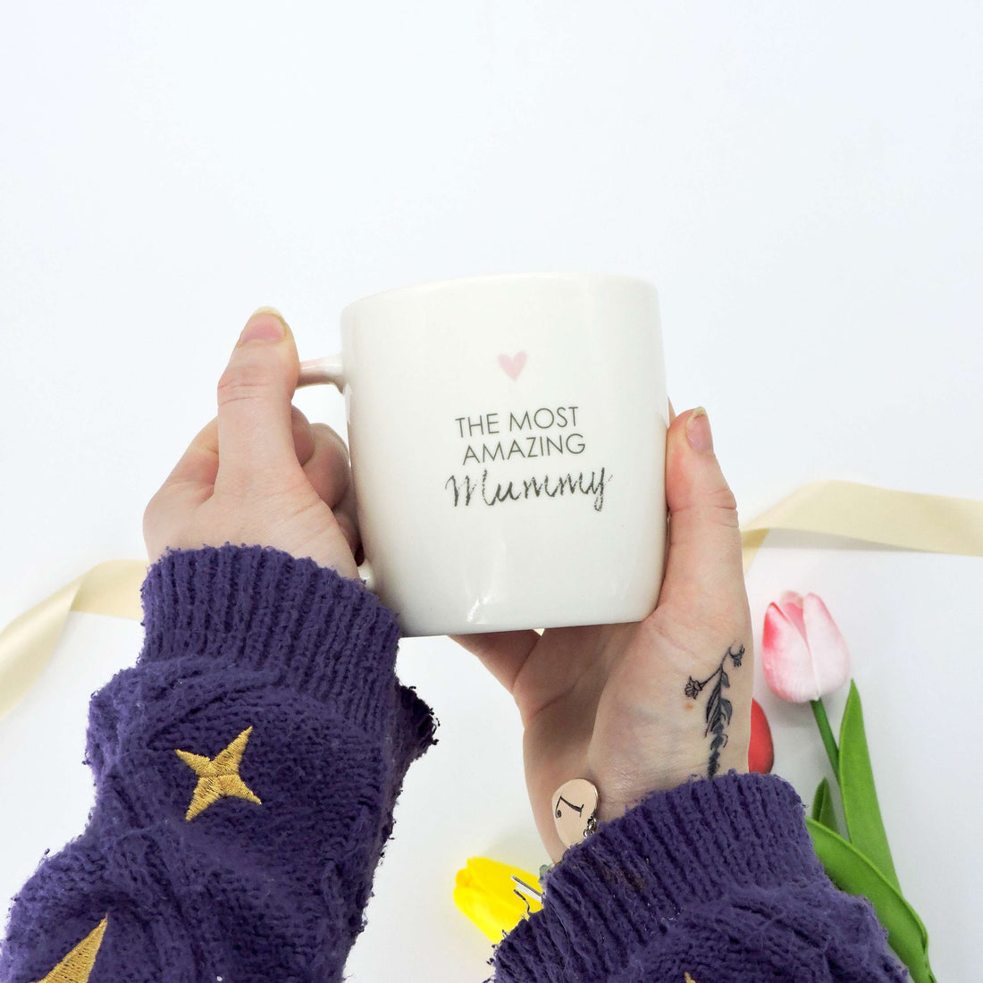 The Most Amazing Mummy Ceramic Mug, held by a person, features a heartwarming message and a soft pink interior, perfect for gifting cherished moments.