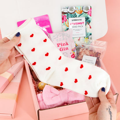 White crew socks with red heart patterns being held up, a cozy addition to a letterbox-friendly pamper gift box.