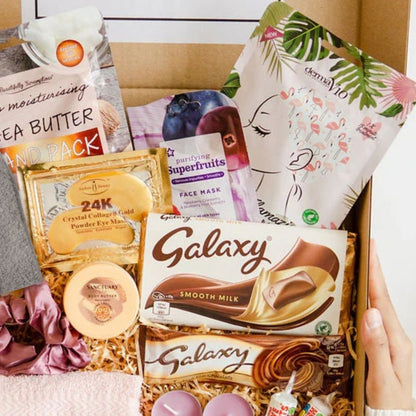 A curated beauty gift box filled with assorted skincare products, including Galaxy Smooth Milk chocolate, a tea butter pack, a face mask packet, and eye cream, arranged in shredded paper inside a cardboard box. A hand is holding the right side of the box. Made by Heavenly boxes. Makes an ideal self care gift for her.