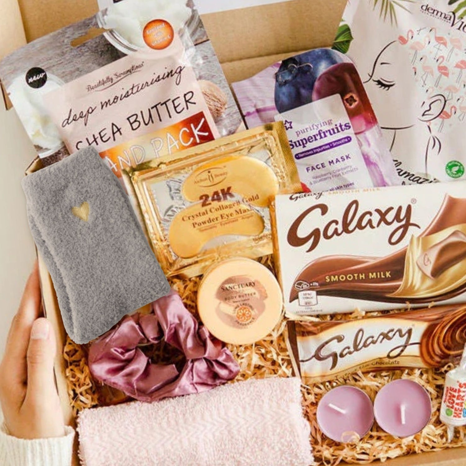 A curated gift box filled with assorted beauty and snack items, including a grey eye mask, a pink satin scrunchie, a round cream makeup compact, Galaxy chocolate boxes, a face mask packet, and two pink candles, arranged in shredded paper inside a cardboard box. Made by Heavenly boxes. Makes an ideal self care gift for her.